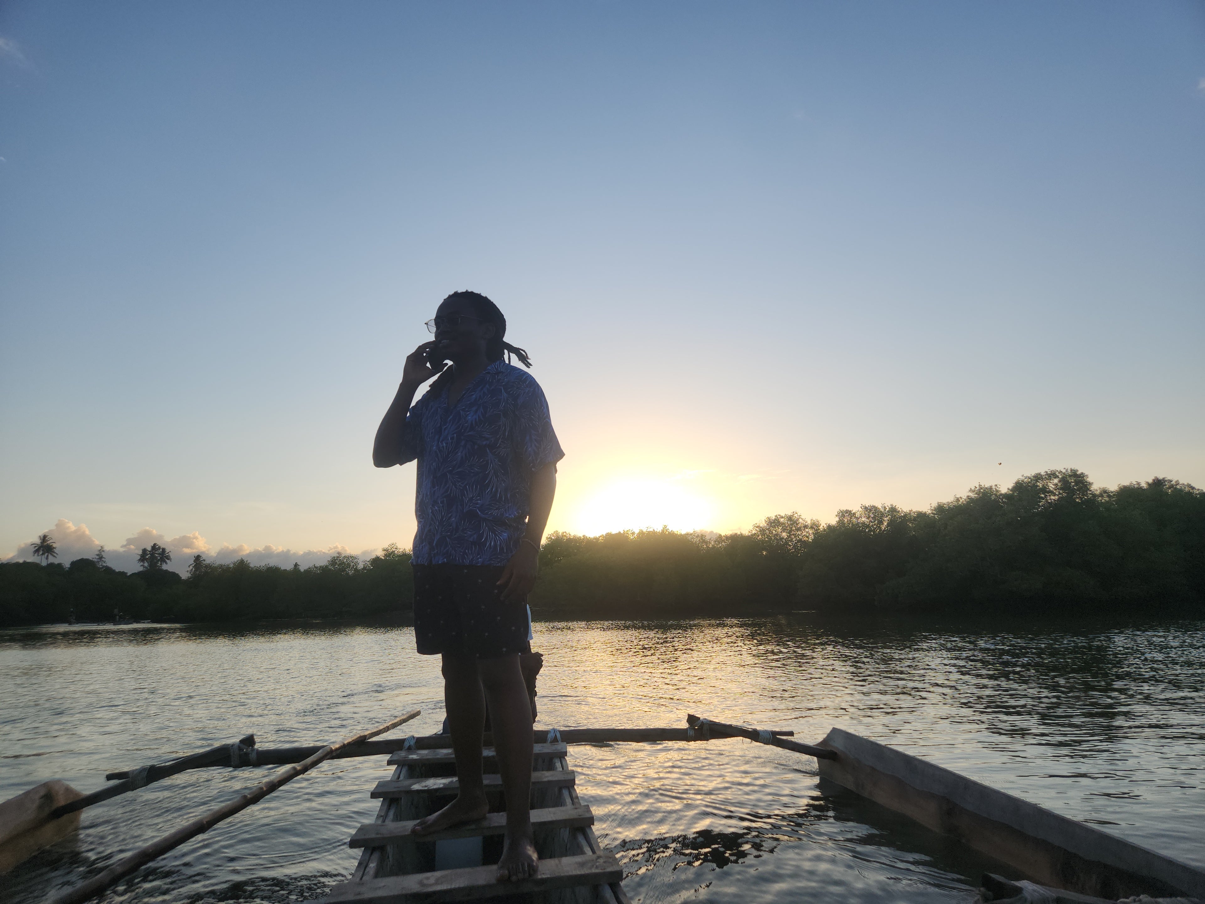 Man on a wooden raft at sunset over water with trees in the background